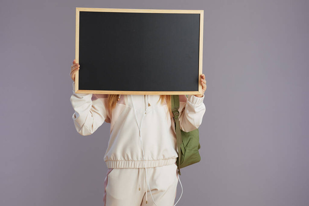 trendy school girl in beige tracksuit with backpack showing blank board isolated on grey. - 写真・画像