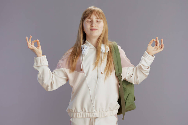 relaxed trendy school girl in beige tracksuit with backpack and headphones isolated on grey. - Zdjęcie, obraz