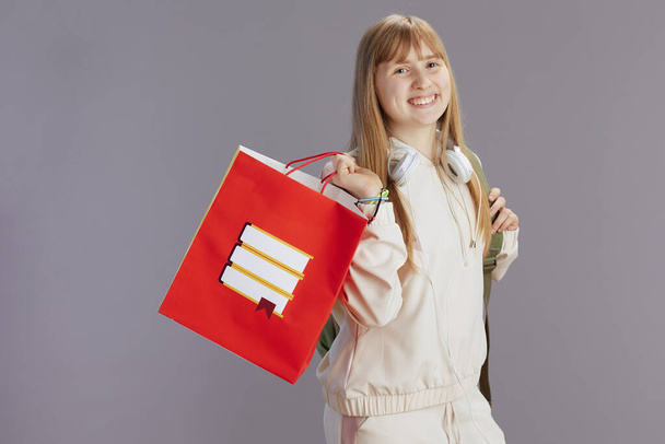 happy modern pupil in beige tracksuit with shopping bag and headphones against grey. - Fotó, kép