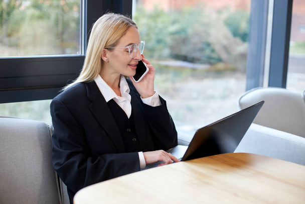 Молода ділова жінка в офіційному костюмі говорить на мобільному телефоні, використовуючи ноутбук в сучасному офісі
 - Фото, зображення