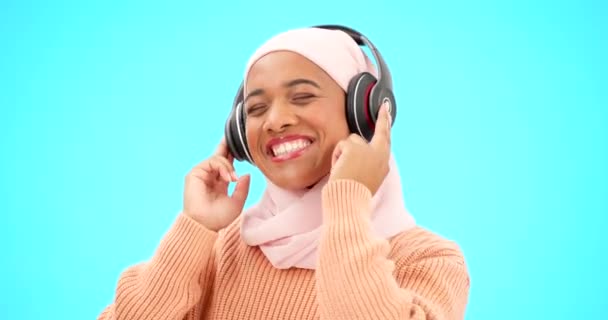 Ευτυχισμένη γυναίκα, ακουστικά και χορός απομονωμένα σε μπλε φόντο ακούγοντας μουσική για γιορτή και τραγούδι. Αστεία, χορός και μουσουλμανισμός, gen z ή hijab άτομο με τεχνολογία streaming στο στούντιο. - Πλάνα, βίντεο