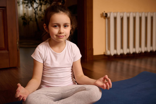 Beautiful European relaxed little kid girl in sports uniform, smiling looking at camera while meditating in lotus position on yoga mat. - Foto, imagen