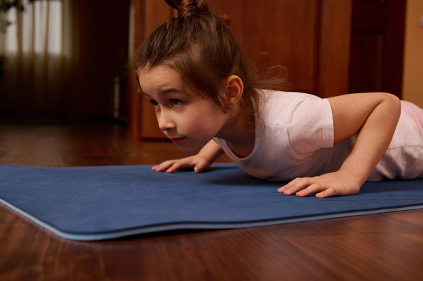 Close-up retrato de uma criança caucasiana 5-6 anos de idade, linda menina em sportswear, fazendo empurrar para cima no tapete de fitness no interior acolhedor casa. Hábitos saudáveis. Bem-estar. Conceito de estilos de vida ativos - Foto, Imagem
