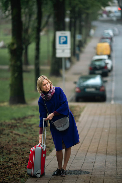 A woman with a suitcase, standing on the sidewalk waiting for a cab.  - Фото, изображение