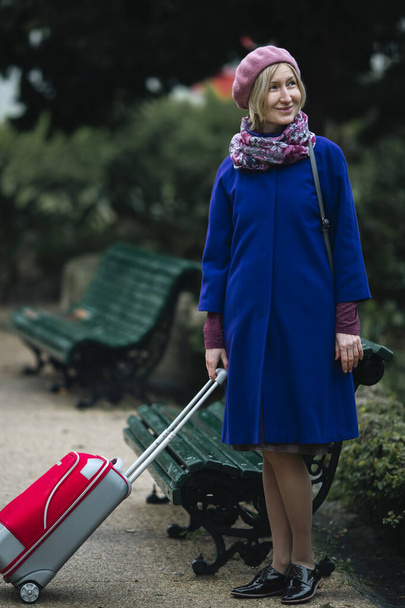 Eine Frau mit einem Koffer steht neben einer Bank im Park. - Foto, Bild