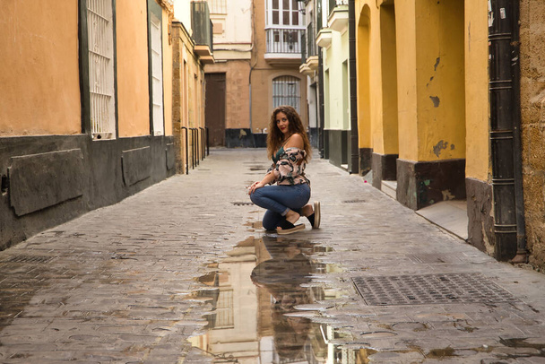 Young and beautiful woman, blonde, with curly hair and blue eyes, wearing floral shirt, green top and jeans, crouched sensually posing on a lonely street, reflected in a puddle. - Photo, Image