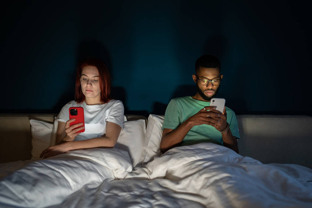 電子機器や家族関係。夕方にベッドに横になっている間にスマートフォンを使用している若い多人種のカップルは、お互いに無視して話していません。インターネット中毒 - 写真・画像
