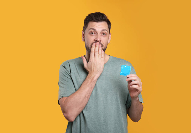 Confused man holding condom on orange background - Foto, Imagen