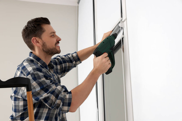 Man with drill installing roller window blind on stepladder indoors - Zdjęcie, obraz