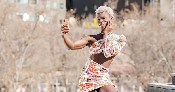 Positiivinen nuori Afrikkalainen Amerikan transsukupuolinen henkilö värikkäässä mekossa ottaen omakuvan älypuhelimella poseeratessaan valokuvaa kadulla - Valokuva, kuva