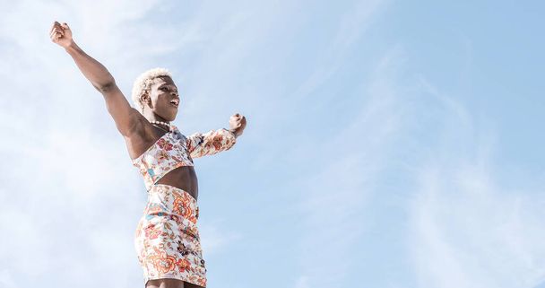 Low angle of carefree young African American transgender person in colorful dress dancing against blue sky and smiling while enjoying freedom on sunny day - Photo, Image