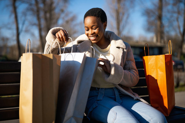Ulkomuotokuva onnellisesta mustasta naisesta. Hän istuu kadulla penkillä ja katselee ostoskassia.. - Valokuva, kuva