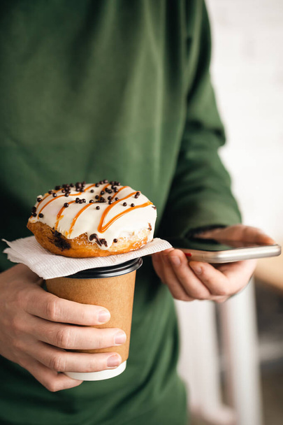 A cup of coffee, a donut and a smartphone in male hands, close-up, appetizing glazed donut. - Photo, image