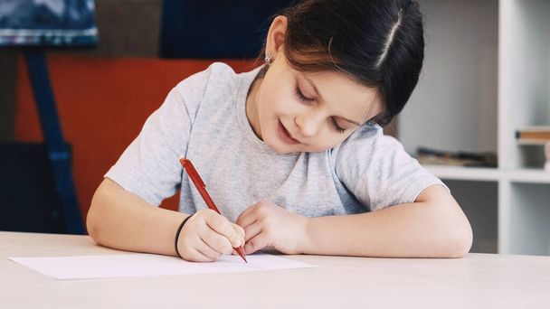 Kids painting. Art hobby. Creative time. Happy girl drawing with pencil on paper sitting table in light room interior. - Photo, image