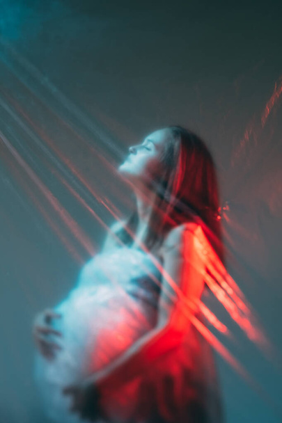 Maternity blessing. Art portrait. Fertility dream. Defocused inspired relaxed pregnant woman silhouette in red blue color light rays. - Foto, imagen