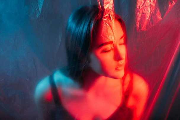 Art portrait. Beauty face. Enhancement rejuvenation. Red blue color light defocused silhouette of relaxed woman with closed eyes behind wrinkled transparent plastic film. - Foto, afbeelding