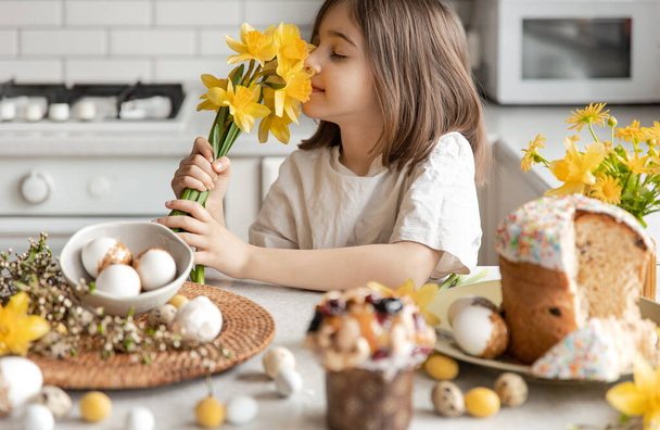 Κοριτσάκι με μπουκέτο από κίτρινα λουλούδια ασφόδελους στην κουζίνα, ψήσιμο και διακοσμητικά αυγά στο τραπέζι, έννοια της προετοιμασίας για το Πάσχα. - Φωτογραφία, εικόνα
