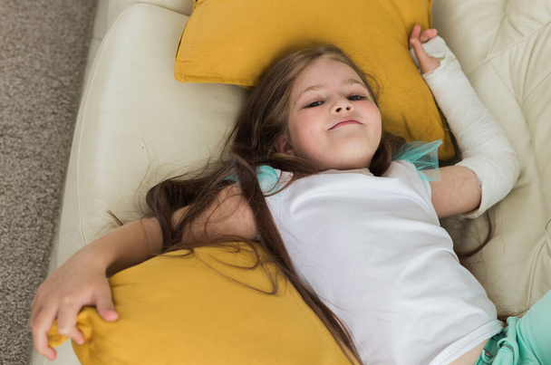 Ребенок со сломанным запястьем или рукой, улыбающийся и веселящийся на диване. Позитивное отношение, выздоровление и ребенок. - Фото, изображение