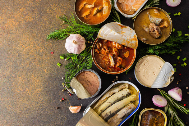 Different open tin cans with canned fish among spices and herbs on a brown background, canned salmon and mackerel, sprat and sardine, tuna and herring and fish pate, top view - Photo, Image