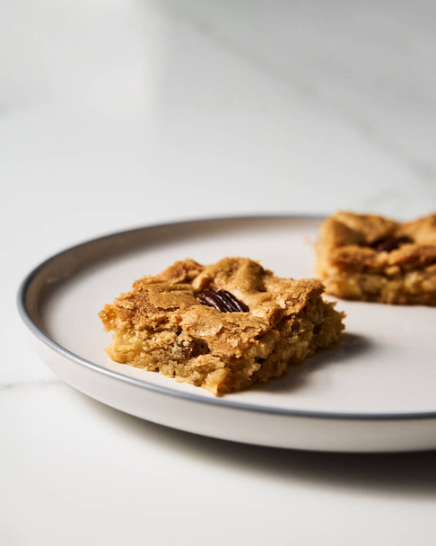 Two squares of Blondie cookies with pecan nuts on white plate. White chocolate sweet and tasty, chewy cookie bars. Close up. High quality photo - Foto, immagini