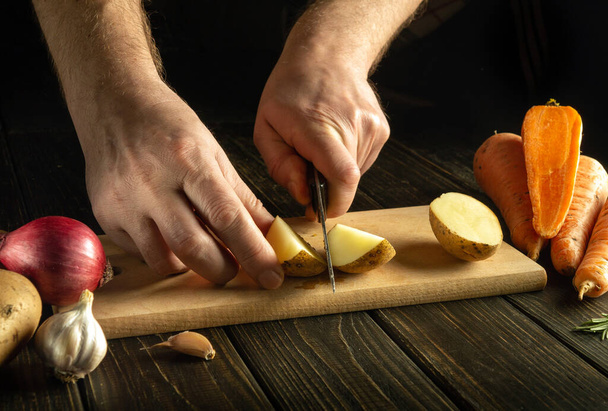 Close-up van de handen van een kok met een mes gesneden rauwe aardappelen op een houten snijplank. Een groenteschotel koken in een keuken van een restaurant. - Foto, afbeelding