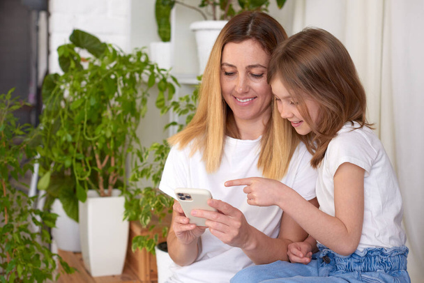 moeder en dochter online winkelen thuis met behulp van smartphone, kind meisje wijst naar mobiele scherm favoriete app - Foto, afbeelding