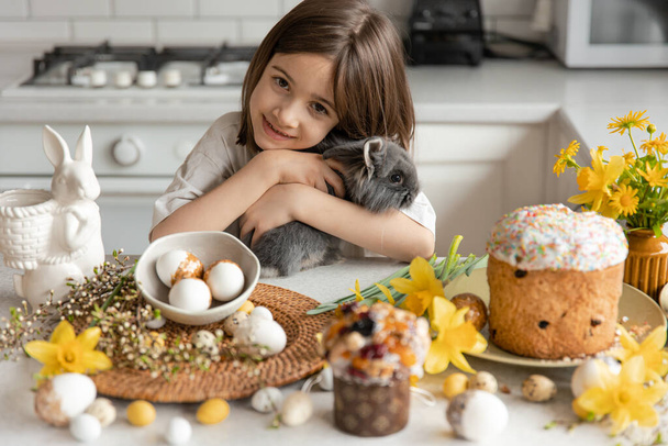イースターエッグ、カップケーキ、花とキッチンの内部にウサギを持つ小さな女の子。イースター休暇のコンセプト. - 写真・画像