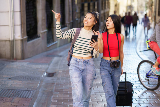 Молодые позитивные азиатские девушки с рюкзаком и чемоданом, гуляющие по улице Гранады и указывающие на достопримечательности во время путешествия - Фото, изображение