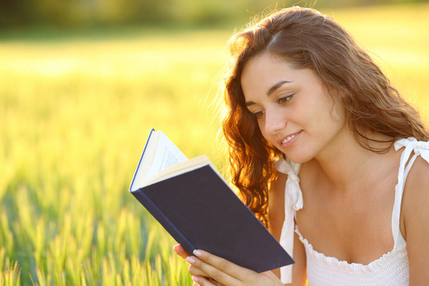 Szczęśliwa kobieta siedząca na polu i czytająca gazetę o zachodzie słońca - Zdjęcie, obraz