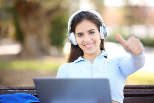 Happy student with laptop and headphone with thumbs up on a bench in a park - Fotoğraf, Görsel