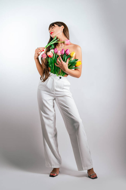 Una chica delgada presiona flores en su pecho desnudo. Hermosa chica con un ramo de tulipanes aislados sobre fondo blanco - Foto, Imagen