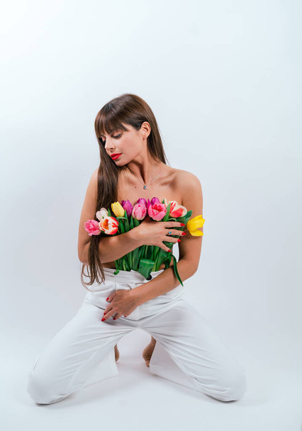 Szczupła dziewczyna przyciska kwiaty do gołej piersi. Piękna dziewczyna z bukietem tulipanów odizolowanych na białym tle - Zdjęcie, obraz