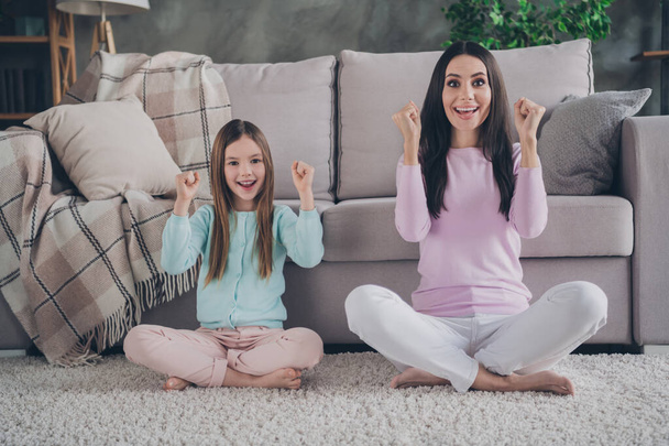 Photo of lucky funky mother daughter wear casual outfits sitting floor rising fists indoors house room. - Фото, изображение