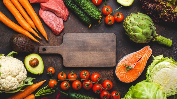 Assorted food raw products: vegetables, beef meat, fish salmon and empty wooden cutting board in centre, dark rustic brown stone table top view. Healthy food background, ingredients for balanced diet - Photo, image