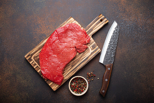 Raw uncooked top round beef steak on wooden cutting board with big kitchen knife and pepper on dark brown rustic stone background top view, cooking meat steak concept - Photo, Image