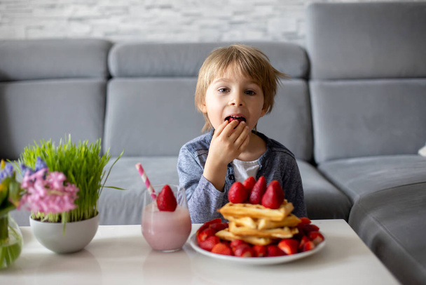 Sweet preschool child, boy, eating belgian waffle with strawberries and chocolate at home and driking smootie - Foto, afbeelding