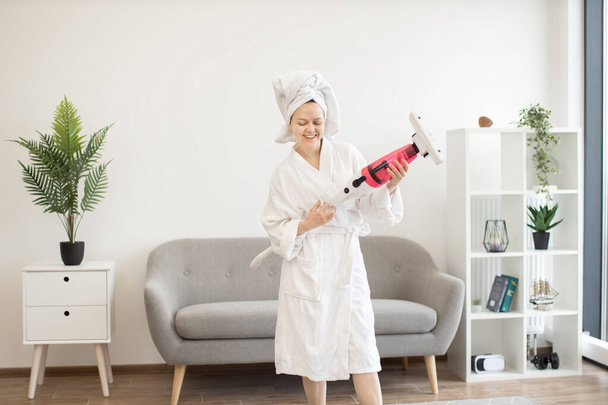 Smiling young adult in dressing gown holding stick vacuum in hands and pretending to be guitar player. Cheerful housewife enjoying modern lifestyle while having fun with cleaning equipment at home. - Fotografie, Obrázek