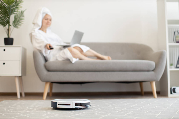 White automatic robot vacuum keeping carpet dust-free while female owner relaxing with portable computer on cozy couch indoors. Wireless tech gadget providing healthy environment in airy apartment. - Φωτογραφία, εικόνα