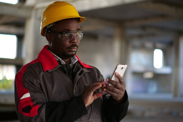 Afrikkalainen amerikkalainen työmies keltaisessa kypärässä älypuhelimella seisoo rakennustyömaalla. Työntekijä omistaa käsissä älypuhelin - Valokuva, kuva