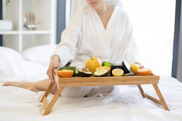 Cropped view of hands of young lady in dressing gown putting tray table with fruits and cosmetic containers on soft bed in bright studio room. Caucasian woman preparing for skin care treatment. - Foto, Imagen