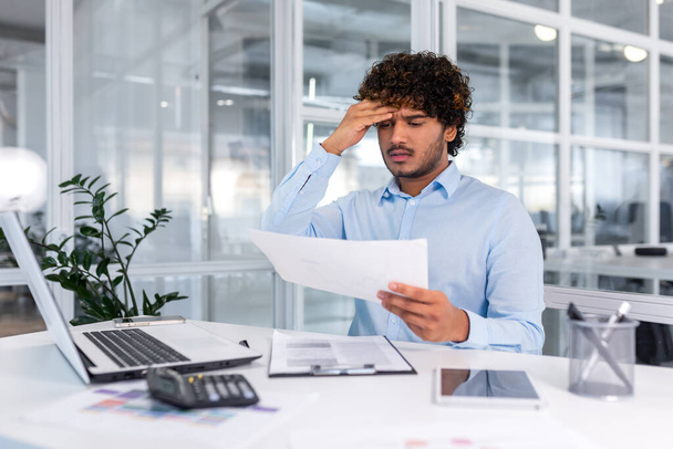 Upset financier looking at sales and revenue chart of company, businessman inside office disappointed with sales results and achievements, Hispanic man sitting at table with documents and laptop. - Foto, Bild