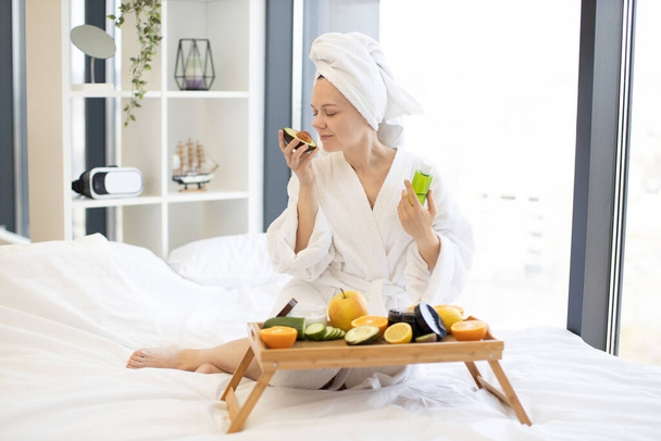 Pretty young woman in after-shower wear smelling fresh avocado while sitting on soft bed in front of tray table with natural skincare remedies. Serene lady pampering herself with spa day at home. - Фото, изображение