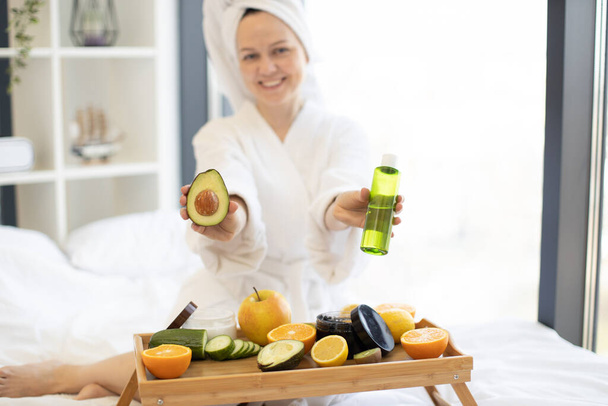 Mujer sonriente en traje después de la ducha presentando aguacate verde en fruta y aceite cosmético mientras está sentada detrás de la bandeja de la cama en casa. Señora alegre recomendando ingredientes naturales en la terapia facial. - Foto, Imagen