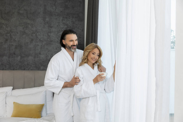 Handsome couple in bathrobe in the bedroom standing near the window curtains - 写真・画像