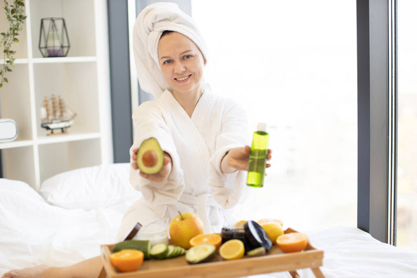 Charming lady in bathrobe smiling with ripe avocado and cosmetic glass vial in hands while resting on bed with fruits and jars on tray table. Pretty adult choosing remedy for gentle skin treatment. - Foto, Imagen