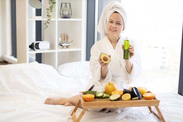 Happy adult female in white robe and towel showing alligator pear with facial oil while enjoying spare time in bedroom during weekend. Joyful lady pampering herself with face care treatment at home. - Foto, Bild