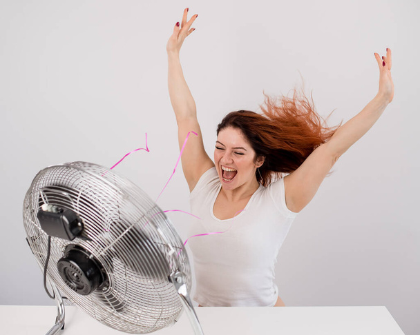 Joyful caucasian woman enjoying the wind blowing from an electric fan on a white background - Fotografie, Obrázek