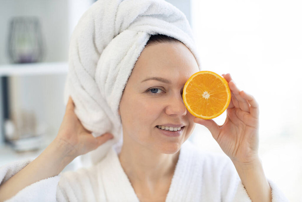 Close-up view of young healthy female wearing soft towel and holding orange fruit piece over her eye on blurred background of bright room. Happy lady achieving natural beauty effect of skin glow. - Foto, Imagem