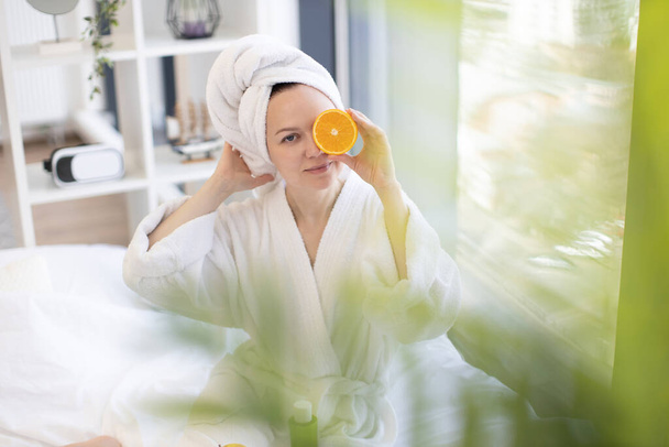 Serene healthy female after bath taking pleasure of fresh citrus fruit aroma while relaxing in peaceful bedroom interior. Charming adult person using rich in vitamin C orange for high-end beauty life. - Foto, imagen