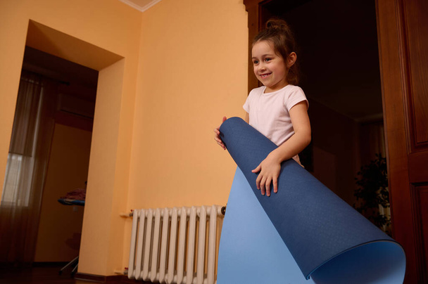 Smiling sporty little girl holding yoga mat while practicing yoga, meditation or doing stretching exercises at cozy homely atmosphere - Photo, Image